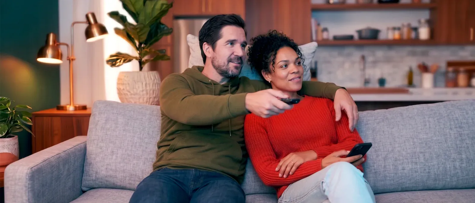 A smiling couple sitting together on a grey sofa in a living room, with the man pointing a remote control toward the television.